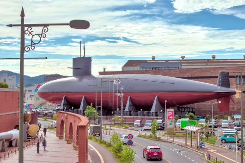 A large submarine sits beside a road and buildings as cars drive by.
