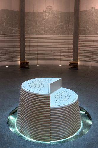 Water runs invisibly over a stone table into a glowing moat inside a stone hall. A relief of the Atomic Bomb Dome is seen on the wall.