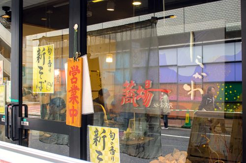 Shirakawa's sign on their glass front with reflections of passing pedestrians in the glass with customers bent over bowl of noodles inside