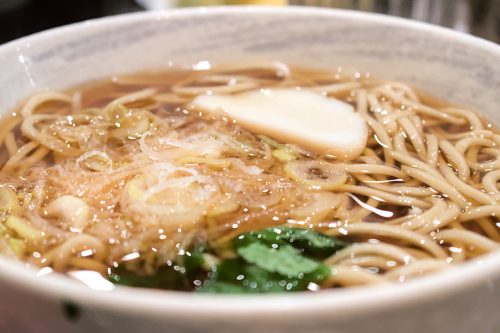 Seen from an angle: a bowl of hot soba in a light-brown, clear broth garnished with mitsuba, negi onions, and fish cake