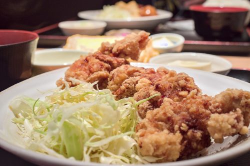 Bumpy and golden brown, pictured is Japanese fried chicken on a plate with a tousle of shredded cabbage and a variety of side dishes in the background