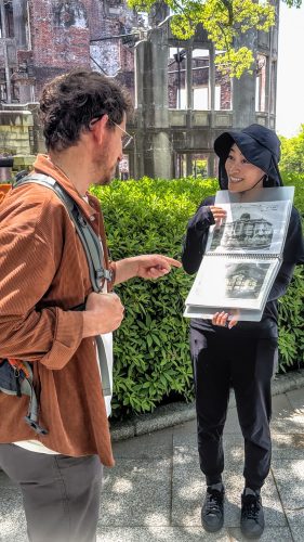 A man points to a picture being held by a woman wearing a sun hat next to the Atomic Bomb Dome