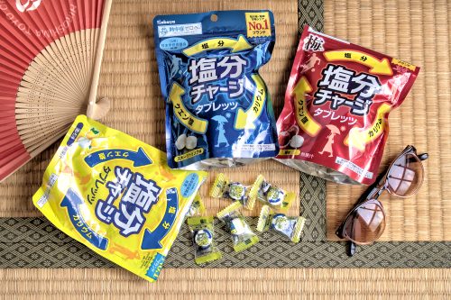 Yellow, blue and red plastic packages with Japanese writing on them. They are on a tatami mat with a pair of sunglasses in one corner and a folding paper fan in the opposite corner