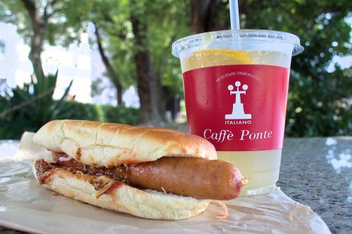 A hotdog and a drink in a plastic cup on a polished stone table under some trees.