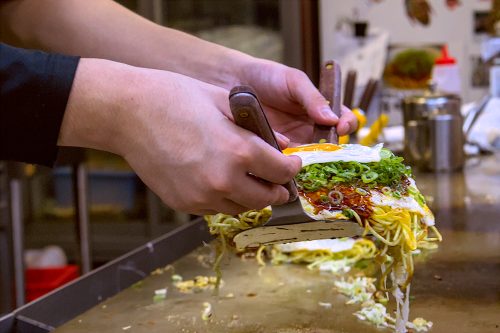 A cook's hands hold up an okonomiyaki on spatulas