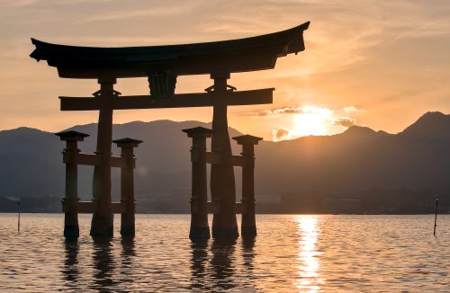 A bright, yellow sunset silhouettes a large Shinto gate that rises out of the sea.