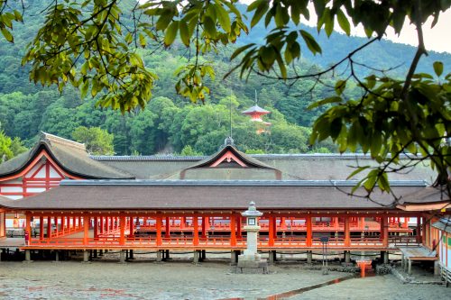 Open air, vermillion hallways rise out of the beachside mud. A green mountain and vermillion pagoda rise in the background.