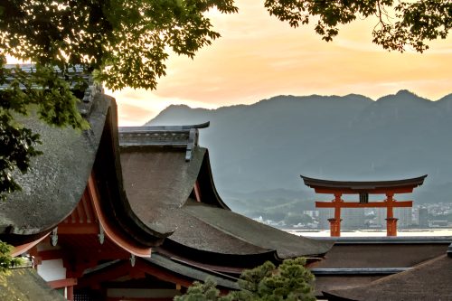 A yellow sunset over mountains with a vermillion Shinto gate rising beyond sloped, Hinoki cypress rooftops.