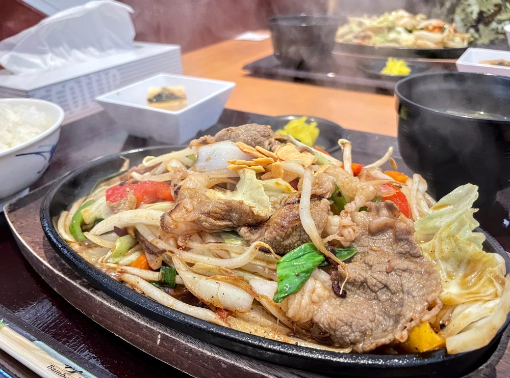A tray with a cast iron plate full of steaming stir-fried vegetables and shaved beef.